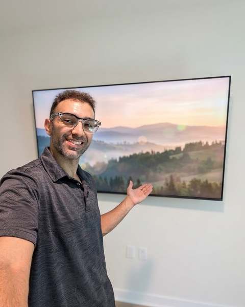 Batu, owner of Batu Home Services, smiling beside a large TV he just mounted on a living room wall
